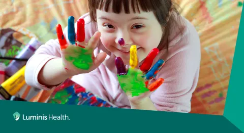 A young child holds up her hands, covered in paint while finger painting.